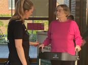 Eine ältere Frau mit Rollator spricht mit einer jüngeren Frau in schwarzer Kleidung vor großen Fenstern in einem hellen Raum.