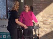 Eine Frau mit schwarzem T-Shirt unterstützt eine ältere Person mit pinkem Pullover, die einen Rollator benutzt, vor einer Backsteinwand.