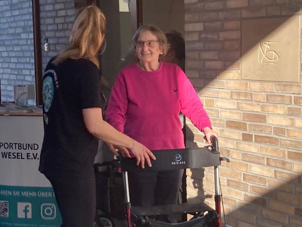 Eine Frau mit schwarzem T-Shirt unterstützt eine ältere Person mit pinkem Pullover, die einen Rollator benutzt, vor einer Backsteinwand.