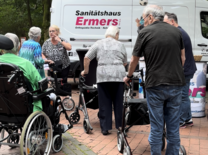Auf dem Weg zum Rollator-Führerschein Ein weißer Kleinbus des Sanitätshauses Ermers, vor dem einige Seniorinnen und Senioren mit ihren Rollatoren und Rollstühlen stehen.