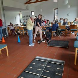 Eine jüngere Frau erklärt einer älteren Dame mit Rollator, wie sie über eine vor ihr auf dem Boden liegende Platte kommt. Im Hintergrund stehen und sitzen weitere ältere Personen und schauen zu.
