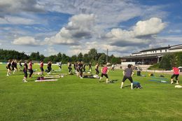 Gruppe von Menschen macht Gymnastik auf einer Wiese vor einem Sportstadion unter bewölktem Himmel.