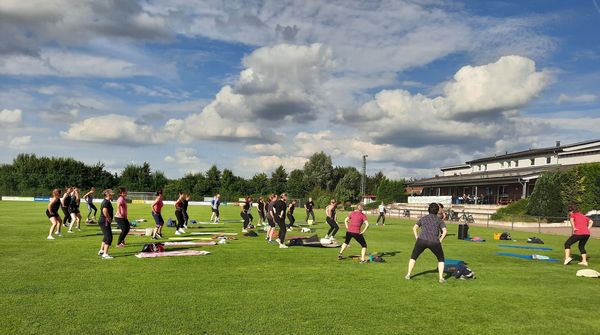 Gruppe von Menschen macht Gymnastik auf einer Wiese vor einem Sportstadion unter bewölktem Himmel.