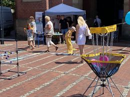 Ein kleines Mädchen im gelben Kleid springt ein Hüpfspiel mit auf dem Boden liegenden Fußabdrücken. Die Mutter steht daneben und schaut ihr zu.