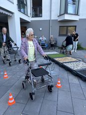 Auf dem Weg zum Rollatorführerschein. Eine ältere Frau mit einem Rollator bewegt sich auf einem gepflasterten Weg, umgeben von orangefarbenen Pylonen. Im Hintergrund sind weitere Personen mit Rollatoren.